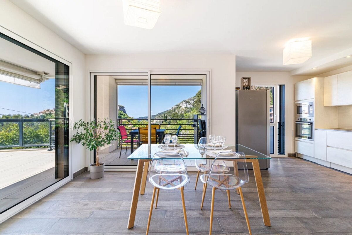 Modern dining room with a glass table and clear chairs, open to a white kitchen and a balcony with beautiful mountain and valley views.