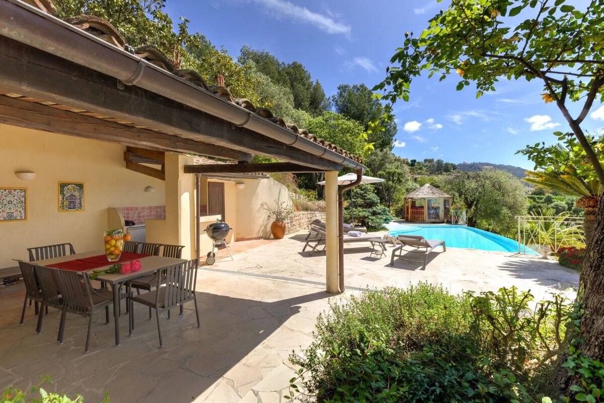A sunny backyard patio features a dining set under an awning, leading to a swimming pool with lounge chairs and lush greenery.
