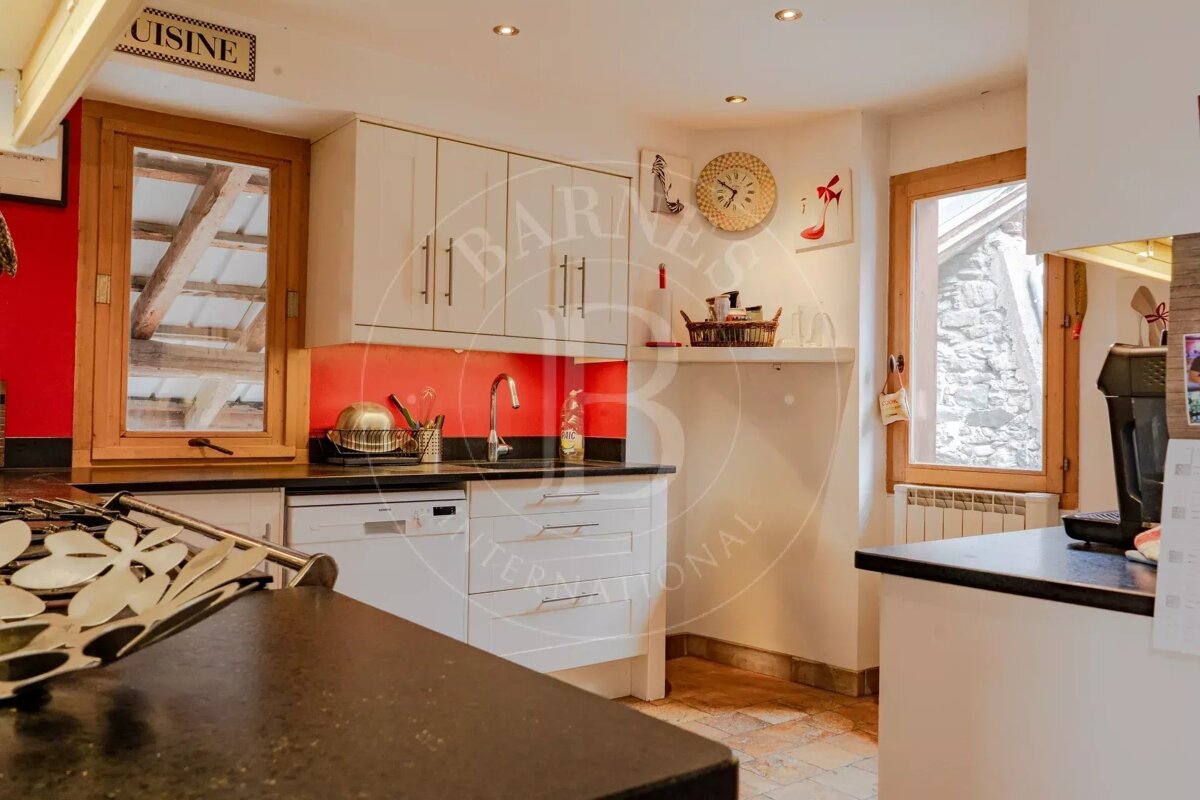A cozy kitchen with white cabinets, a red backsplash, black countertops, and wooden-framed windows offering views of rustic architecture.