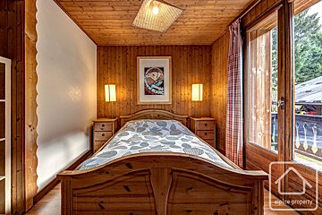 A cozy wooden bedroom with a double bed, framed poster, side tables with lamps, and large glass doors leading outside.
