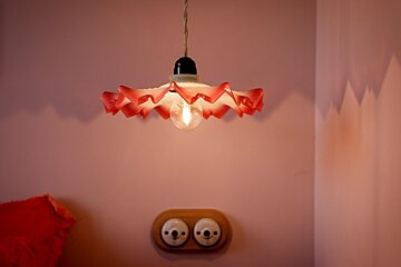 A retro red & white ruffled lamp with a glowing bulb hangs above two ceramic outlets on a pink wall, casting shadows.