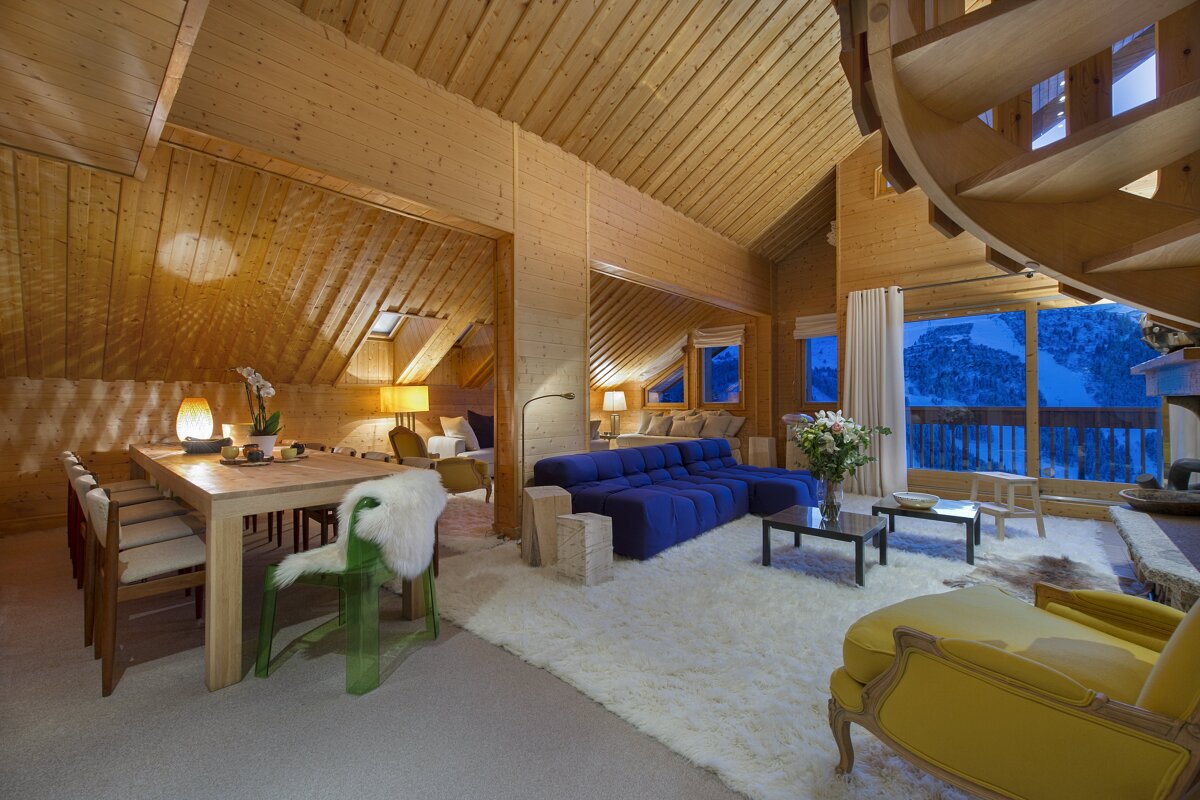 Warm, wood-clad chalet interior with a dining area, vibrant blue sofa, cozy white rugs, a spiral staircase, and a panoramic view of snowy mountains.