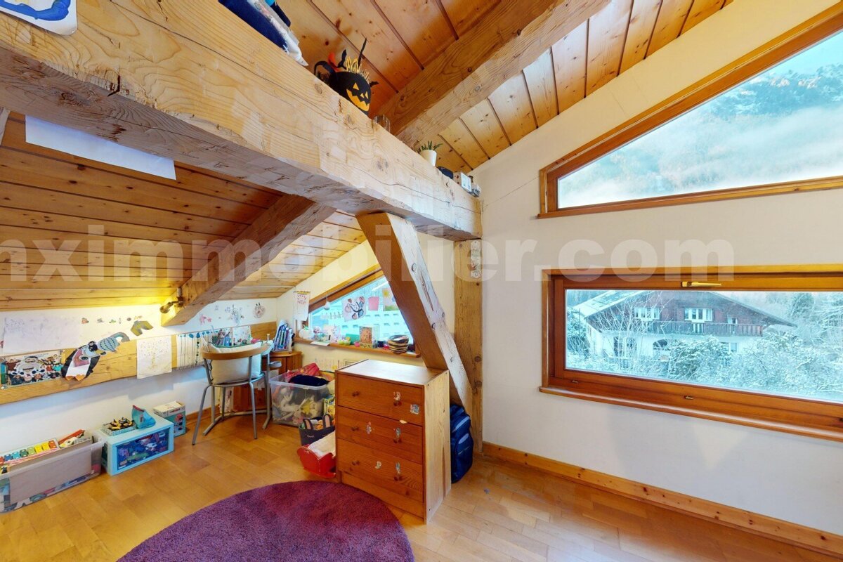 Warm, rustic attic room with exposed wooden beams and sloped ceilings. Features a child's play/study corner and large windows with a snowy outdoor view.