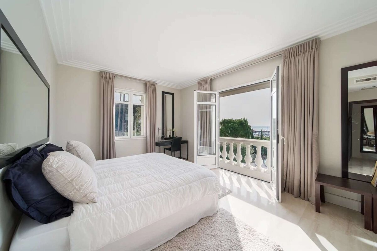 Bright bedroom with a white bed, open balcony, and a scenic view of the sea and coastal trees. Features neutral tones.