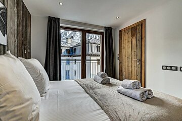 Modern bedroom with a neatly made bed, fresh towels, and a balcony view of snowy town buildings and mountains.
