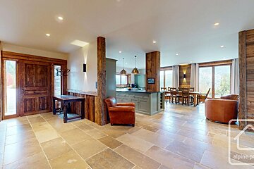 Spacious, rustic open-plan living area with stone floors, wood paneling, a modern kitchen, dining space, leather armchairs, and large windows.