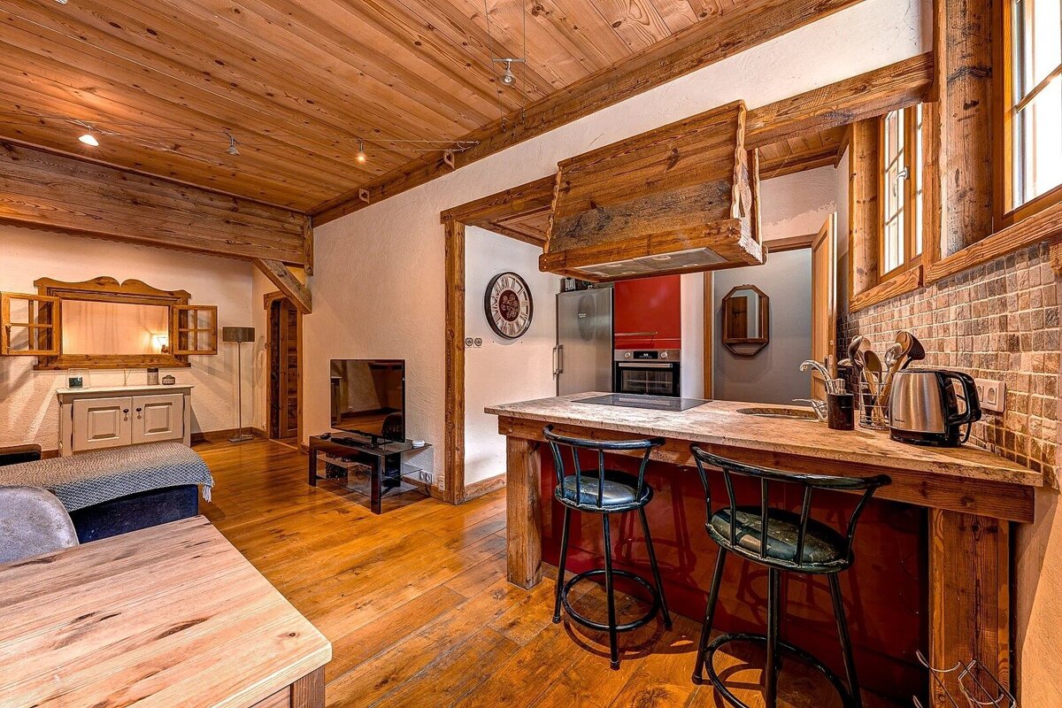 A warm, rustic open-plan living and kitchen area with wooden ceilings, floors, and beams. Features a kitchen bar, modern appliances, and a cozy lounge.