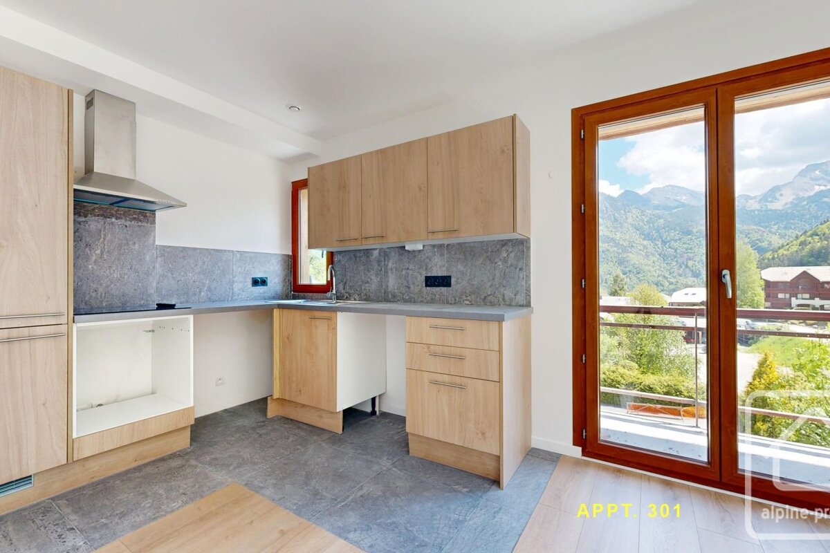 Modern kitchen with light wood cabinets, grey backsplash & flooring, and a large window overlooking lush mountains. Appliances are missing.