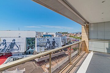 Balcony view overlooking the Cannes Film Festival venue, featuring large billboards and a bright blue sky.