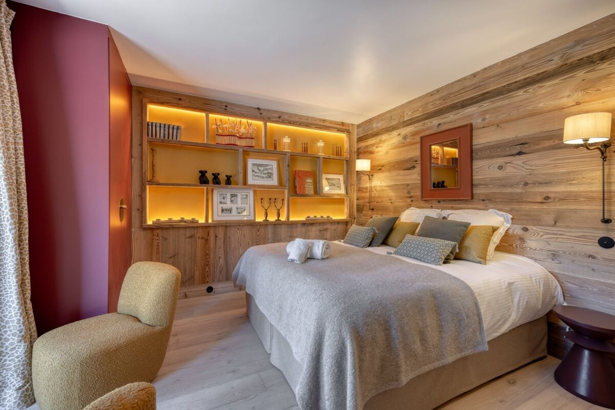 A cozy, rustic bedroom with light wood paneling, an illuminated built-in shelf, a large bed with grey bedding, and a maroon accent wall.