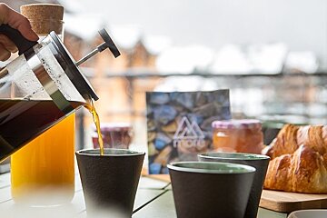 Coffee being poured from a French press into a mug during an outdoor breakfast with croissants and orange juice.