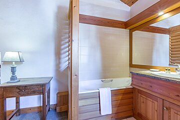 A cozy, rustic bathroom with wood paneling, tiled bath, wooden vanity, mirror, and a small table with a lamp, creating a warm ambiance.