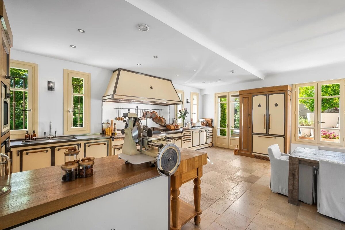 A spacious, elegant kitchen featuring cream-colored appliances, a large range hood, butcher block island with a slicer, tiled floor, and sunny windows.