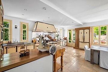 A spacious, light-filled, rustic-style kitchen with cream cabinets, a large range hood, tiled floor, and a vintage meat slicer on a wooden island.