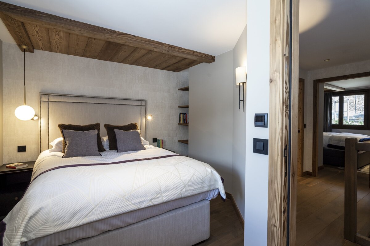 A chic bedroom features a large bed, textured headboard, wood-beamed ceiling, and modern lighting. Neutral tones create a cozy, stylish ambiance.