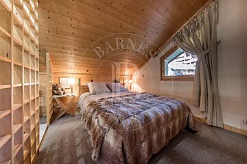 Cozy attic bedroom with warm wood paneling and sloped ceiling, featuring a large bed with a furry throw, a window, and built-in shelves.