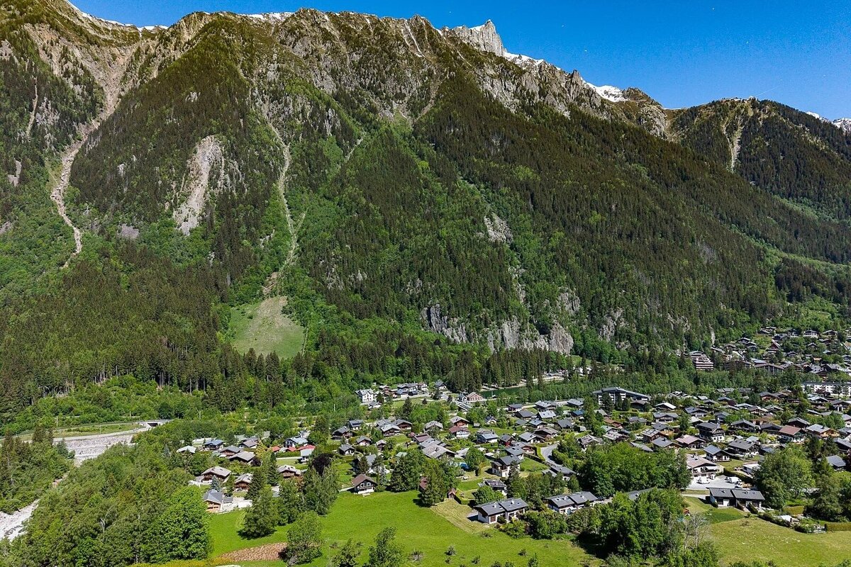 Scenic aerial view of a valley village nestled at the foot of majestic, tree-covered mountains under a clear blue sky.
