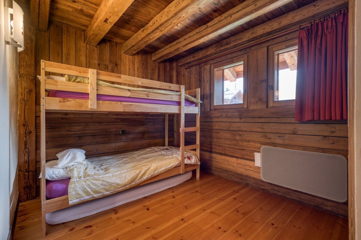 A rustic wooden room features bunk beds, a wooden floor, and ceiling beams. Two windows, one with a red curtain, offer natural light.