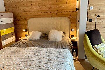 Cozy bedroom with wood paneling, a bed with fluffy pillows and a knit blanket, a colorful dresser, and a yellow armchair.