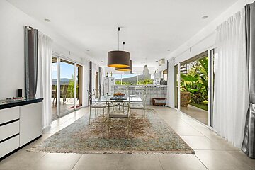 Bright, open-plan dining and kitchen area with glass doors leading to patios and green mountain views. Features a glass dining table & large rug.