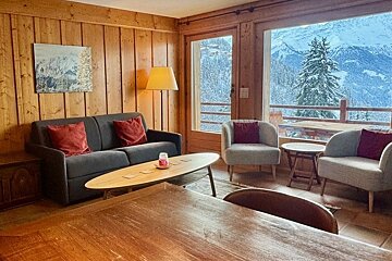 Cozy chalet living room with wood paneling, a sofa, and armchairs looking out at a snowy mountain landscape.