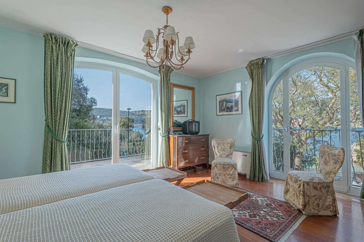 A light blue bedroom with twin beds, a balcony offering a bay view, green curtains, chandelier, dresser with TV, and floral chairs.