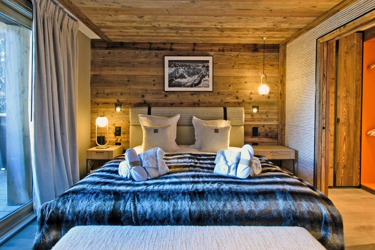 A warm, modern chalet bedroom with wooden walls/ceiling, a plush bed with a faux fur throw, slippers, and a balcony view. Orange closet inside.