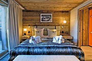 A warm, modern chalet bedroom with wooden walls/ceiling, a plush bed with a faux fur throw, slippers, and a balcony view. Orange closet inside.