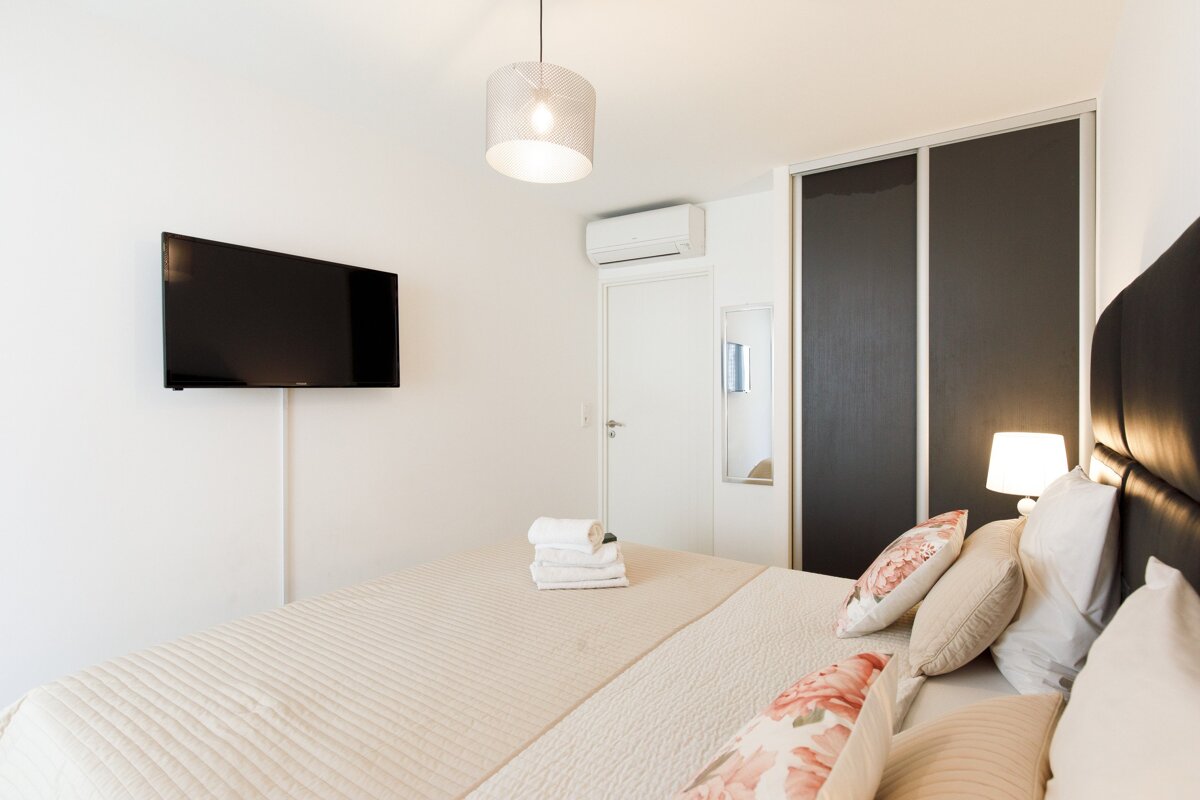 A bright, modern bedroom features a large bed with neutral bedding & floral pillows, a wall-mounted TV, AC, and a dark built-in wardrobe.