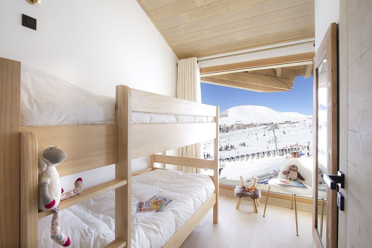 A modern bedroom features light wood bunk beds with white linen and a plush sheep toy, overlooking a stunning snow-covered mountain ski resort.