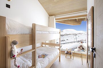 A modern bedroom features light wood bunk beds with white linen and a plush sheep toy, overlooking a stunning snow-covered mountain ski resort.