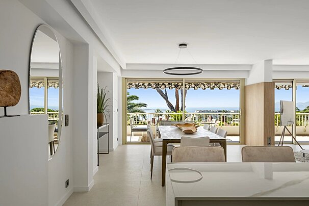 A bright, modern dining room with a long table and sleek chandelier, leading to a balcony with panoramic sea and coastal views.