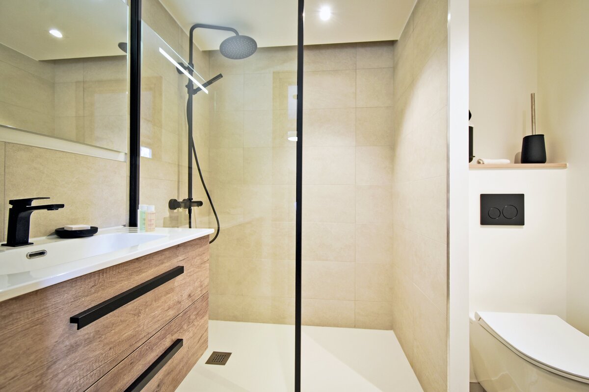 A modern bathroom features a walk-in shower with black fixtures, a wooden vanity with a black faucet, and a toilet, all set against light beige tiles.