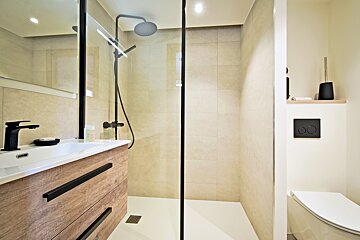 A modern bathroom features a walk-in shower with black fixtures, a wooden vanity with a black faucet, and a toilet, all set against light beige tiles.