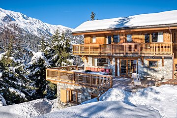 A multi-level wooden chalet with balconies, nestled in deep snow with evergreen trees, backed by majestic snow-capped mountains under a bright blue sky.