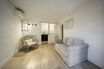 A bright, white living room features a light gray sofa, a wicker chair, and wooden floors. A small kitchen area is visible in the background.