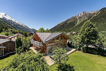 Charming wooden chalet with a green lawn and deck, nestled in a valley with lush trees and majestic snow-capped mountains under a clear blue sky.