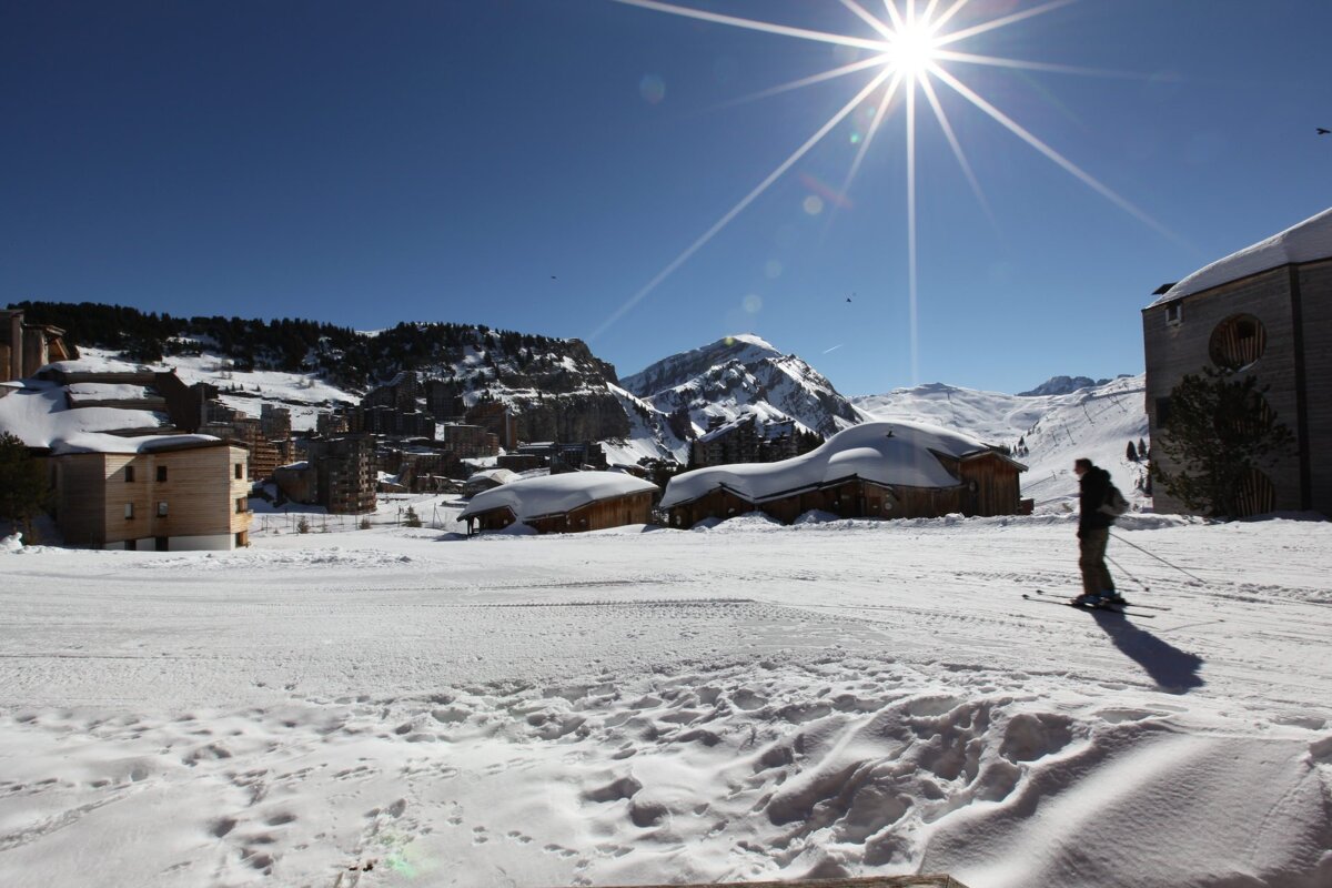Sunny day at a snow-covered mountain ski resort. A skier traverses the foreground with village buildings, majestic peaks, and a bright sun in the blue sky.