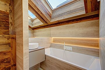 Modern bathroom with a white tub, vanity, light stone walls, rustic wooden beams, skylights, and warm accent lighting.