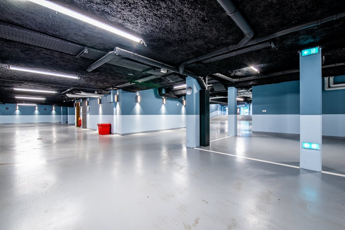 An empty, bright, modern parking garage with blue and white walls, a dark ceiling, exposed pipes, and a red emergency box.