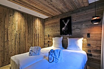 A cozy chalet-style bedroom features rustic wooden walls and ceiling, twin beds with white linen, and a deer print. Slippers/robes are on beds.