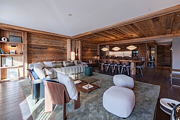 A luxurious, modern chalet living room with rustic wood-paneled walls and ceiling, comfortable seating, and a dining area in the background.