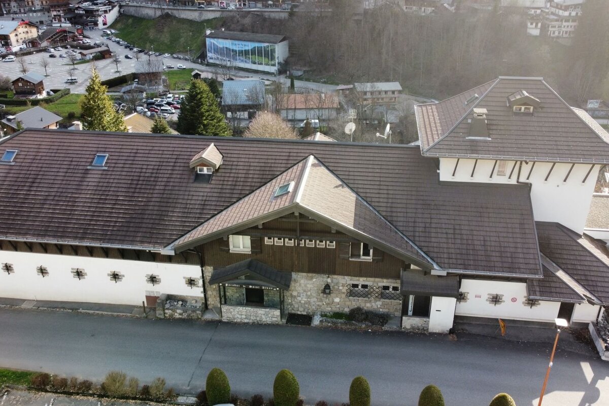 An aerial view of a large building with a multi-gabled brown tiled roof in a mountainous village, with roads, cars, and other buildings visible.