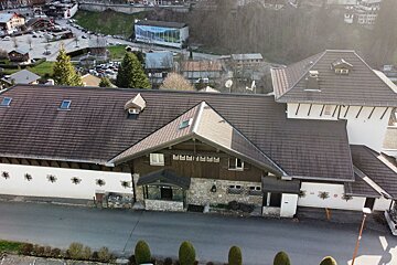 An aerial view of a large building with a multi-gabled brown tiled roof in a mountainous village, with roads, cars, and other buildings visible.