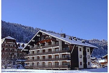 Large chalet-style building covered in snow, with snowy mountains and pine trees in the background, under a bright blue sky.