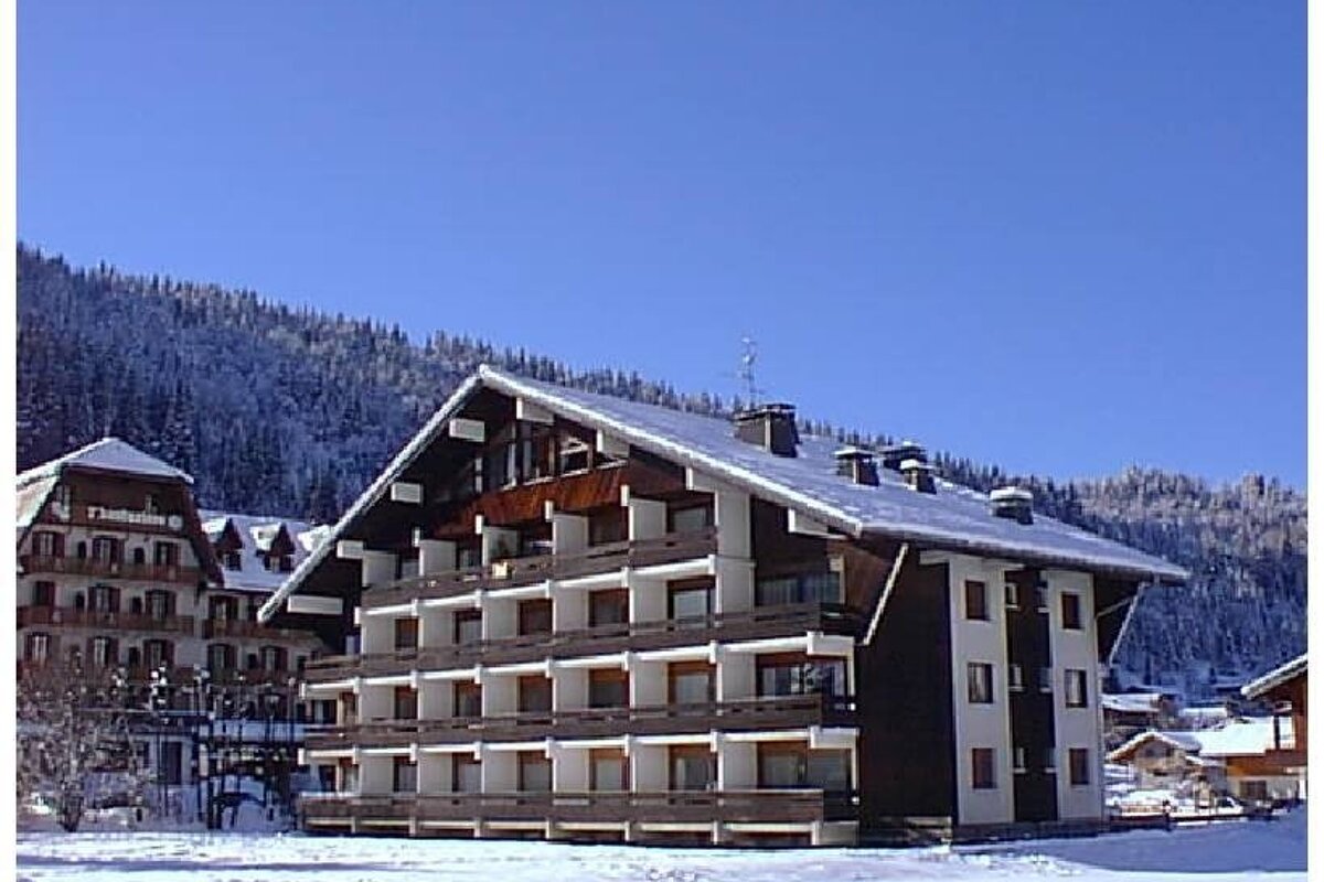 Alpine chalet-style buildings with snow-covered roofs in a winter mountain resort under a bright blue sky.