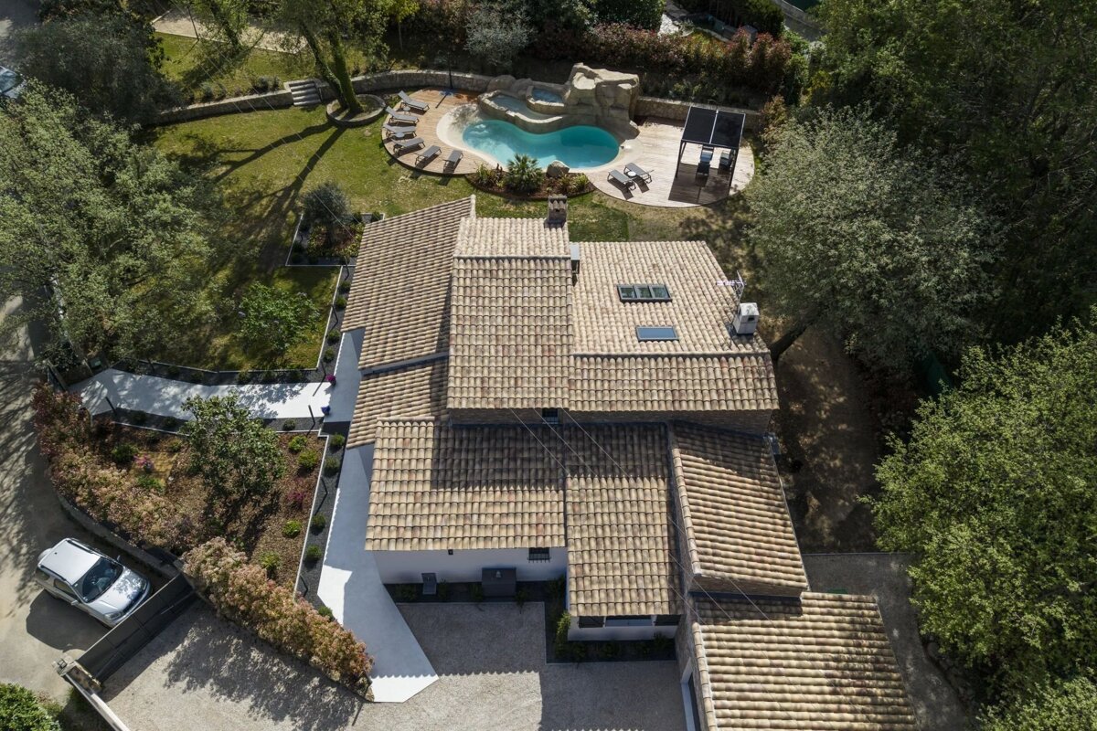 Aerial view of a house with tiled roofs, featuring a backyard with a lagoon-style swimming pool, lounge area, and lush trees.