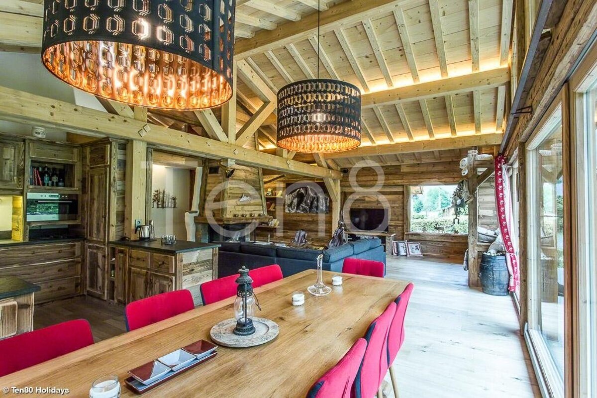 Rustic wooden chalet interior featuring a dining table with red chairs and large pendant lights under an exposed beam ceiling.