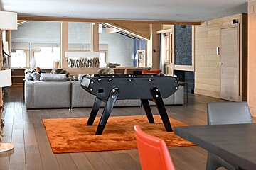 A modern living room features a black foosball table on an orange rug, a grey sofa, wooden walls, and large windows.
