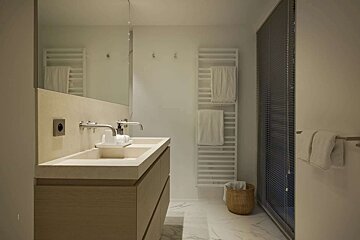 A modern, minimalist bathroom with a double vanity, large mirror, towel warmer, and a glass shower with blinds. Features neutral tones and a marble floor.
